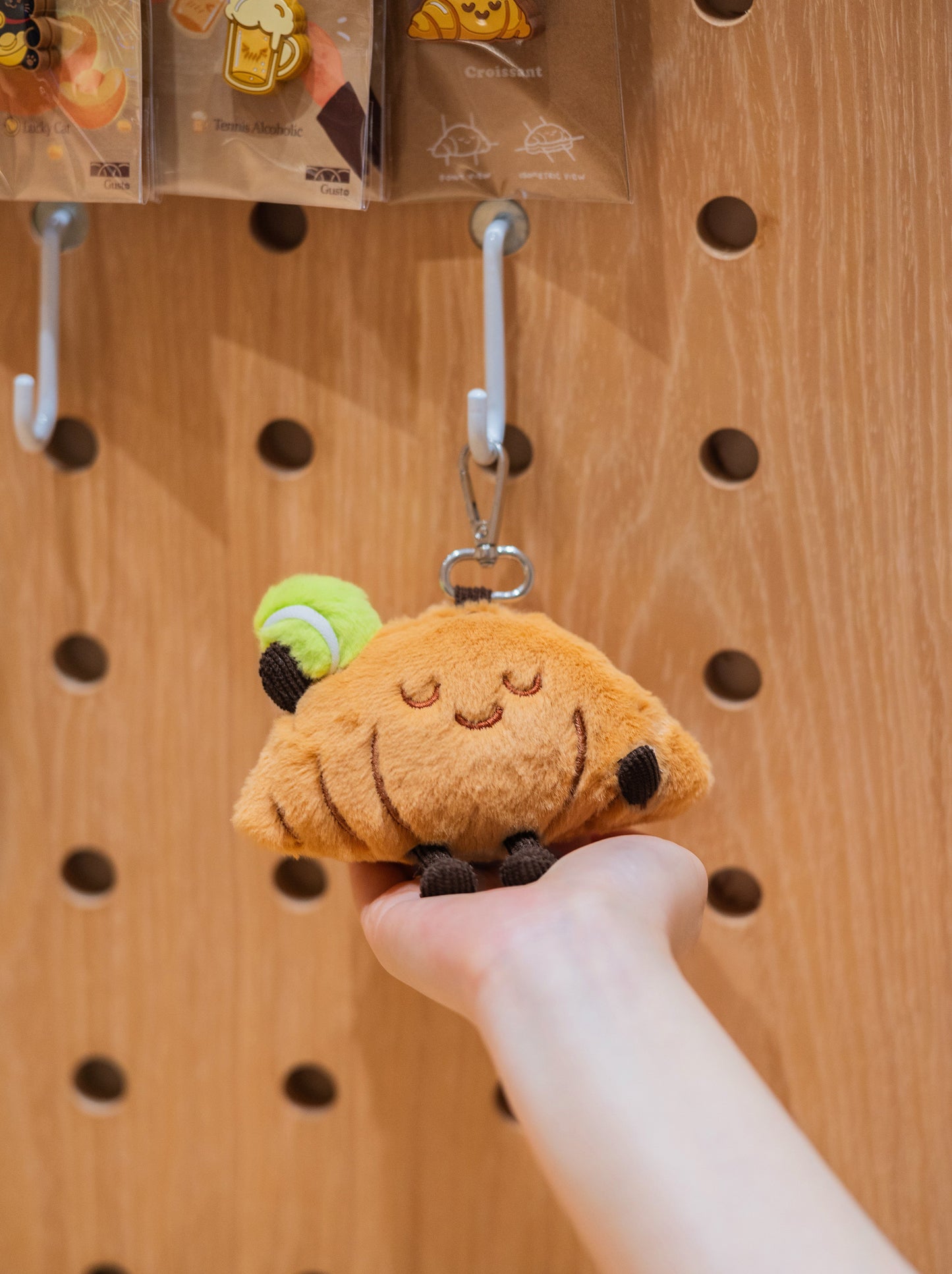 Croissant plush toy held in one hand with tennis merchandise in the background