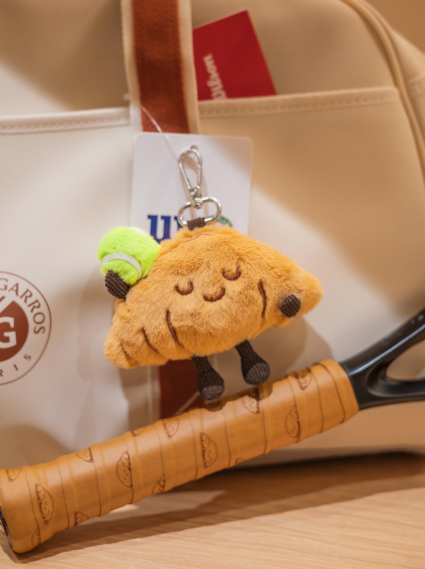 Croissant plush toy with tennis racket and professional tennis bag in product photo