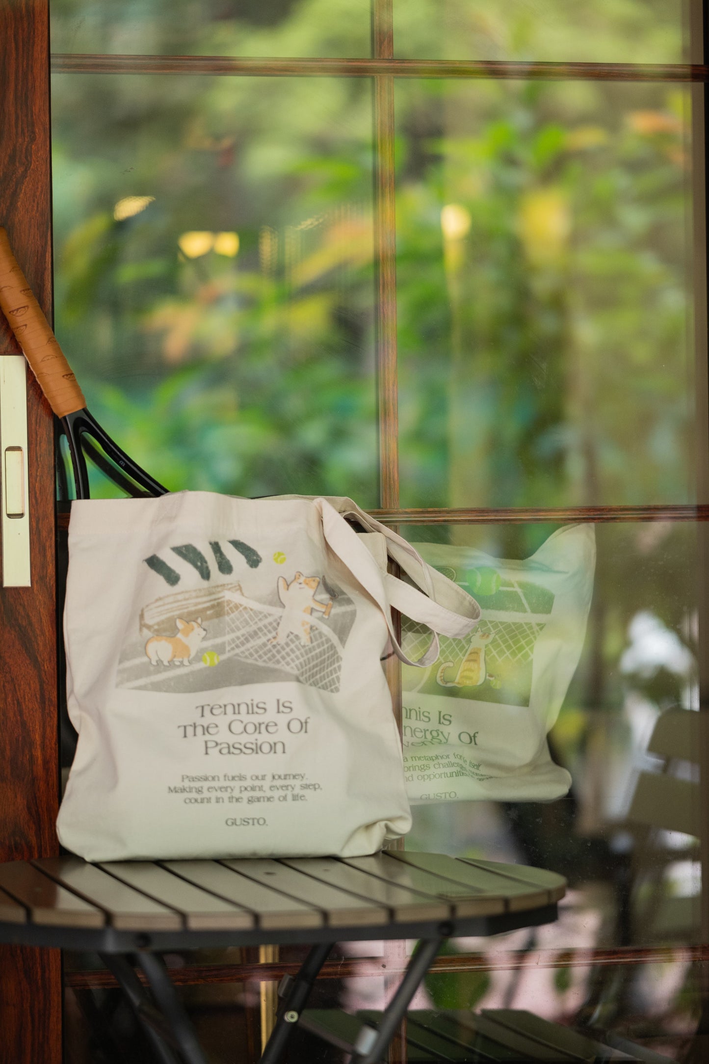 Gray tennis canvas tote bag with corgi design, placed on a café table with a tennis racket inside