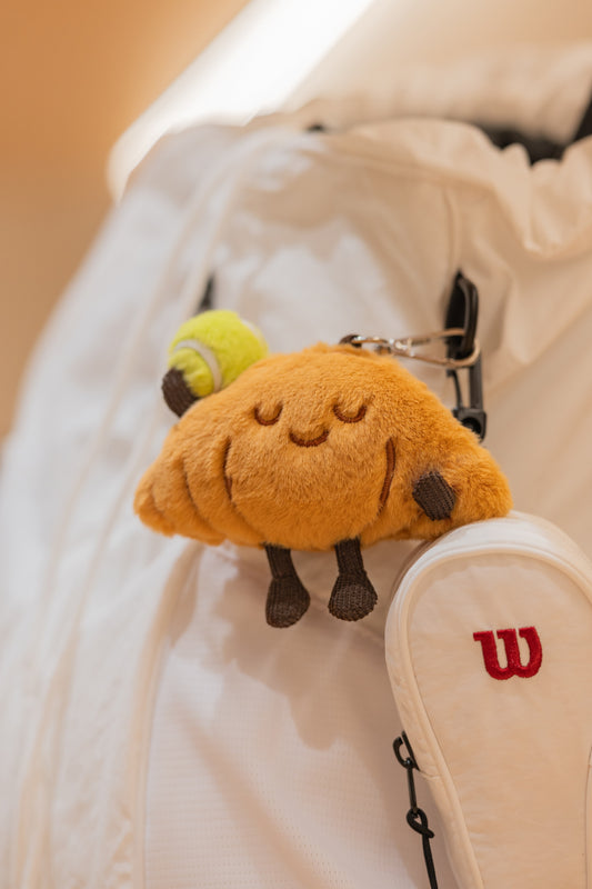 Croissant plush tennis toy hanging as a decoration on a white tennis bag