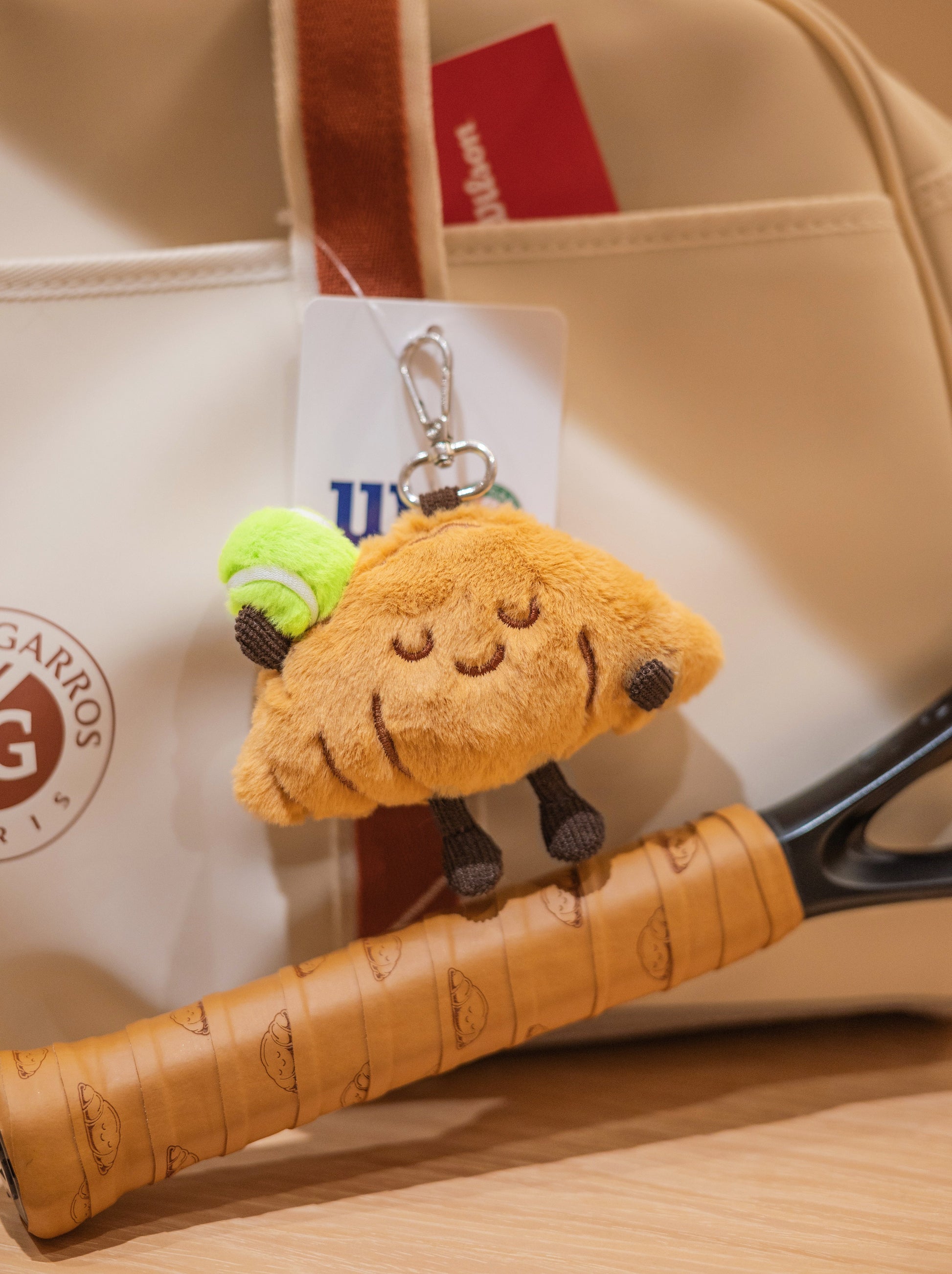 Croissant plush toy with tennis racket and professional tennis bag in product photo