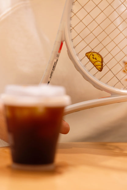 Croissant shaped tennis racket dampener photographed in café lifestyle scene, bakery series accessory
