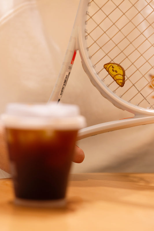 Croissant shaped tennis racket dampener photographed in café lifestyle scene, bakery series accessory