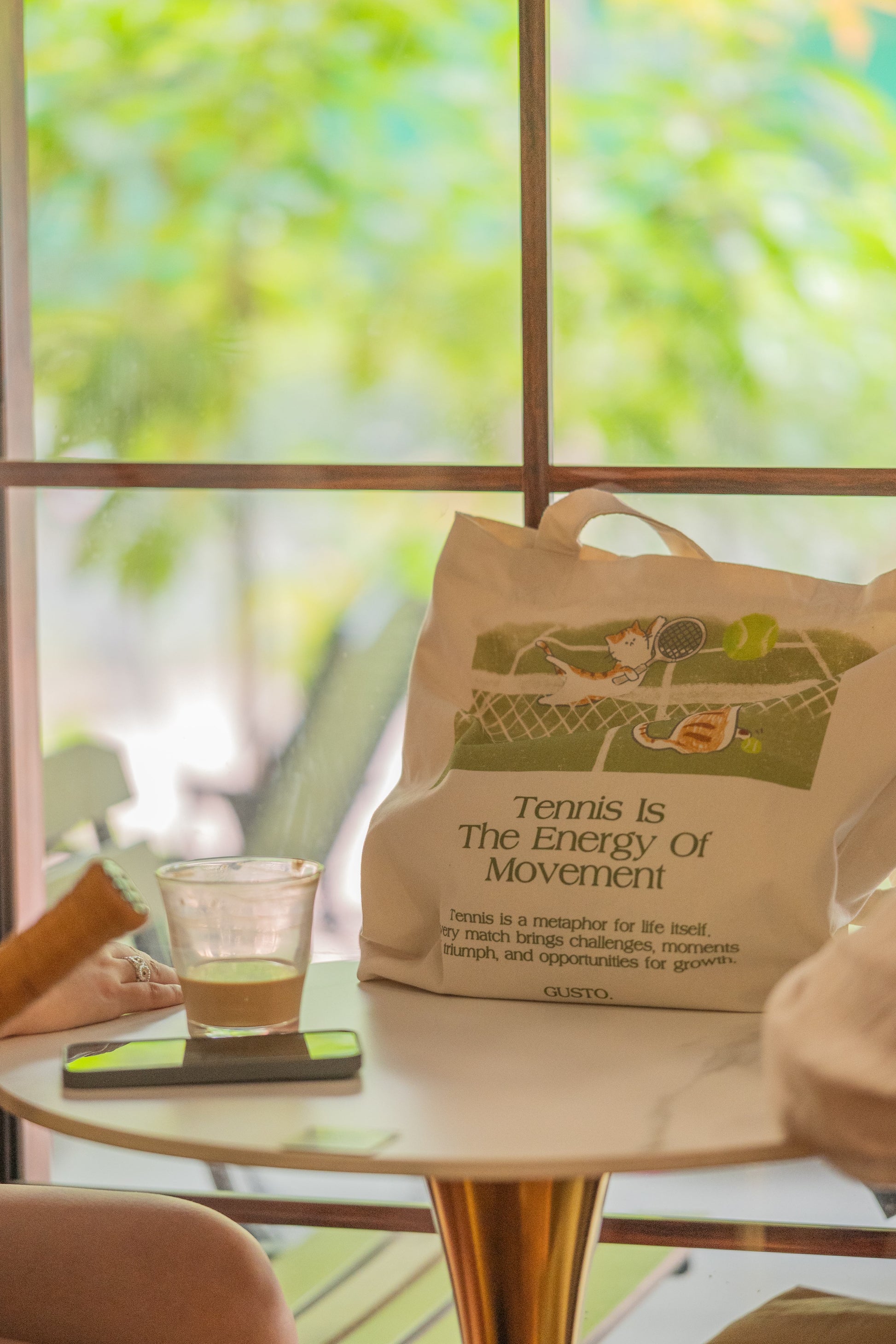 Green tennis canvas tote bag with cat illustration, photographed in a café with leafy trees outside the window