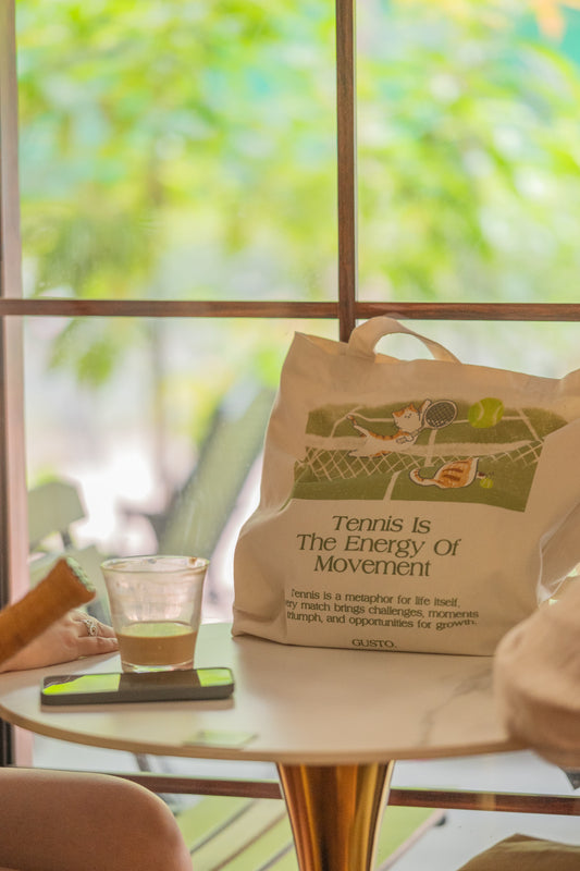Green tennis canvas tote bag with cat illustration, photographed in a café with leafy trees outside the window